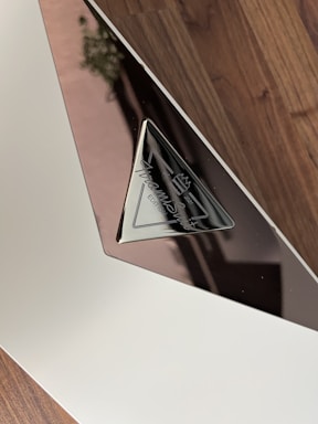 A close-up view of a reflective plaque with a triangular metal section. The plaque features engraved text which appears to be a logo or signature. The background consists of a wooden surface.