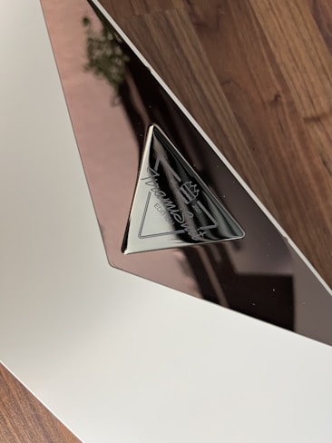 A close-up view of a reflective plaque with a triangular metal section. The plaque features engraved text which appears to be a logo or signature. The background consists of a wooden surface.