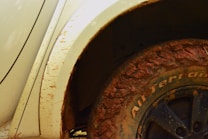 A close-up of a vehicle's wheel covered in mud. The tire has visible tread patterns, and some of the vehicle's body is also stained with dirt. The tire reads 'All Terrain' on the side.