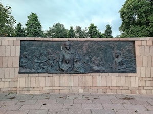 A bronze relief mural depicting a group of people engaged in various activities, including conversations and gestures, set against a stone wall. The mural is framed by stone paving and surrounded by lush green trees under a cloudy sky.