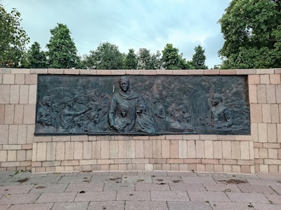 A bronze relief mural depicting a group of people engaged in various activities, including conversations and gestures, set against a stone wall. The mural is framed by stone paving and surrounded by lush green trees under a cloudy sky.