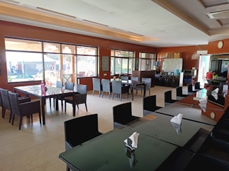 A spacious and modern dining area with several dark-colored tables and wicker-style chairs. Large windows provide ample natural light, and there are decorative elements such as plants and wall art. In the background, a counter with a refrigerator and other kitchen appliances is visible.
