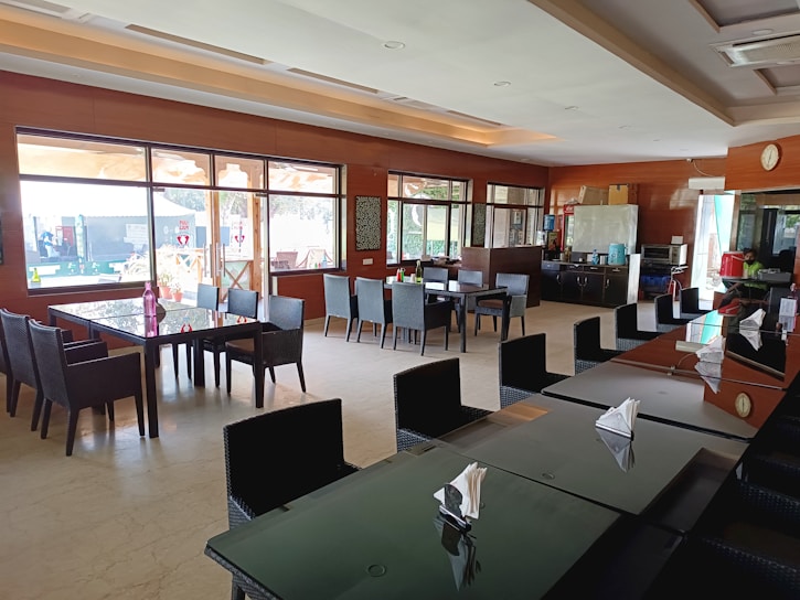 A spacious and modern dining area with several dark-colored tables and wicker-style chairs. Large windows provide ample natural light, and there are decorative elements such as plants and wall art. In the background, a counter with a refrigerator and other kitchen appliances is visible.