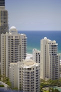 Several tall, modern buildings with domed tops are situated near a coastline. The ocean is visible in the background, and the buildings have large windows and balconies. Palm trees and greenery surround the buildings.