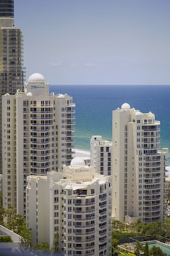 Several tall, modern buildings with domed tops are situated near a coastline. The ocean is visible in the background, and the buildings have large windows and balconies. Palm trees and greenery surround the buildings.
