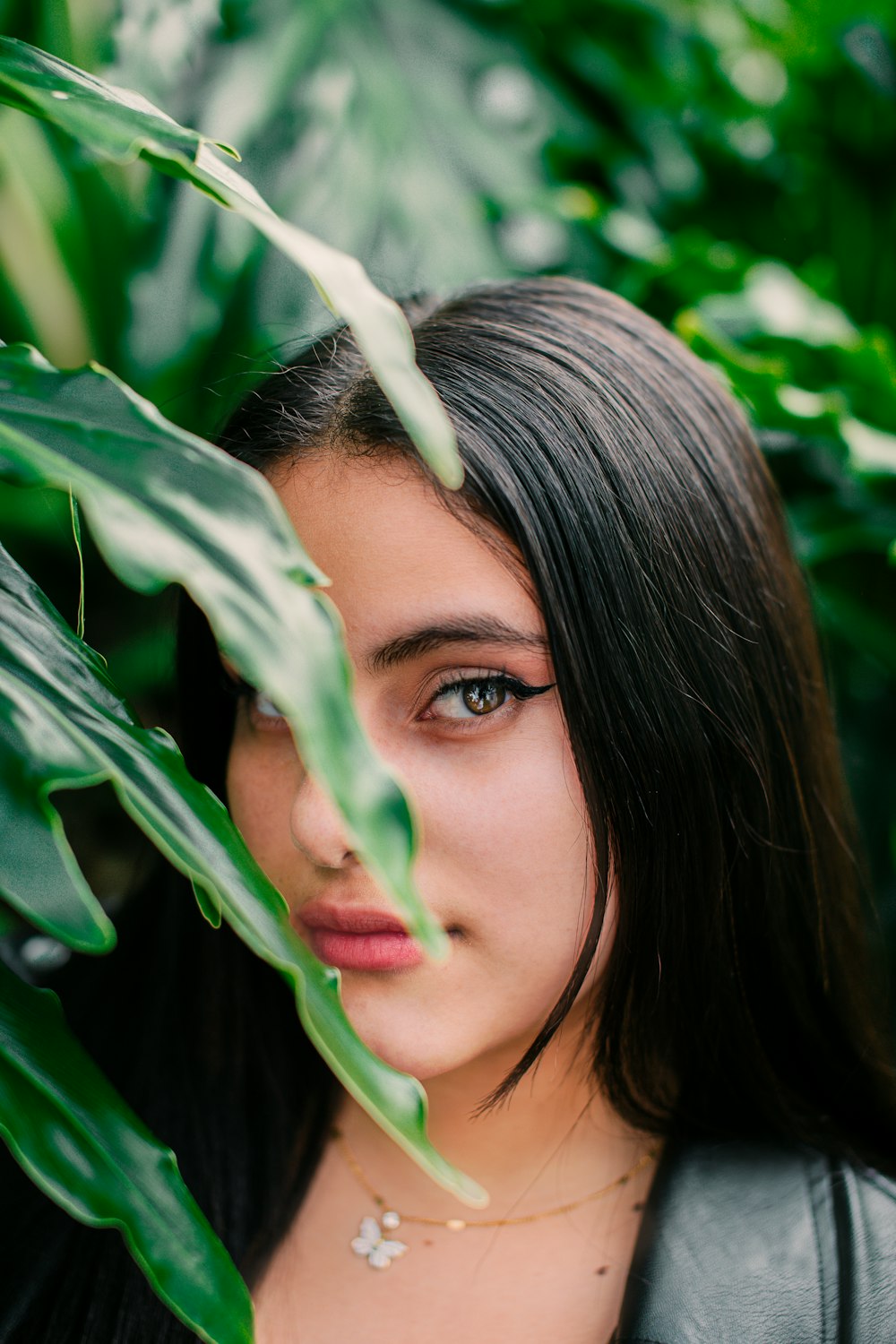 a woman with long black hair standing in front of green leaves