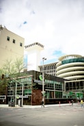The image captures an urban scene featuring a modern building with glass windows and a prominent sign that reads 'NUMERICA Credit Union'. There is a large, cylindrical structure with a unique architectural design in the background. The street is relatively empty with just a few pedestrians and some minimal traffic. A few small trees are visible along the sidewalk, and there is a blend of modern and slightly older architecture.