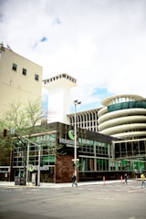 The image captures an urban scene featuring a modern building with glass windows and a prominent sign that reads 'NUMERICA Credit Union'. There is a large, cylindrical structure with a unique architectural design in the background. The street is relatively empty with just a few pedestrians and some minimal traffic. A few small trees are visible along the sidewalk, and there is a blend of modern and slightly older architecture.