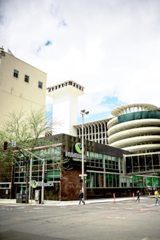 The image captures an urban scene featuring a modern building with glass windows and a prominent sign that reads 'NUMERICA Credit Union'. There is a large, cylindrical structure with a unique architectural design in the background. The street is relatively empty with just a few pedestrians and some minimal traffic. A few small trees are visible along the sidewalk, and there is a blend of modern and slightly older architecture.