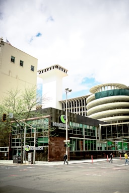 The image captures an urban scene featuring a modern building with glass windows and a prominent sign that reads 'NUMERICA Credit Union'. There is a large, cylindrical structure with a unique architectural design in the background. The street is relatively empty with just a few pedestrians and some minimal traffic. A few small trees are visible along the sidewalk, and there is a blend of modern and slightly older architecture.