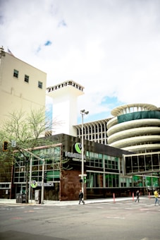 The image captures an urban scene featuring a modern building with glass windows and a prominent sign that reads 'NUMERICA Credit Union'. There is a large, cylindrical structure with a unique architectural design in the background. The street is relatively empty with just a few pedestrians and some minimal traffic. A few small trees are visible along the sidewalk, and there is a blend of modern and slightly older architecture.