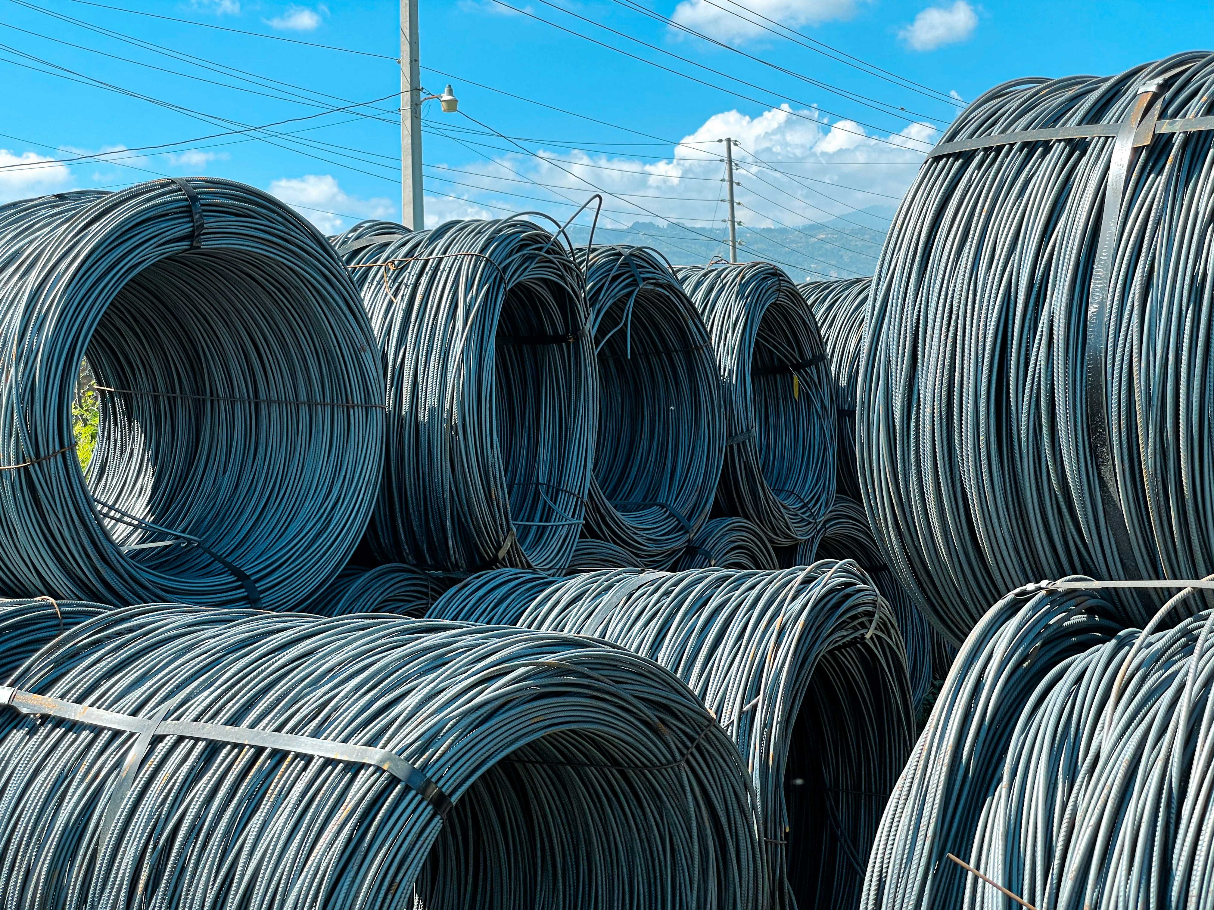 Large coils of steel cables are stacked together outdoors. The cables are neatly wound and secured with metal bands. Power lines and a clear blue sky with scattered clouds are visible in the background.