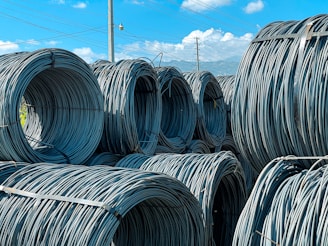 Large coils of steel cables are stacked together outdoors. The cables are neatly wound and secured with metal bands. Power lines and a clear blue sky with scattered clouds are visible in the background.