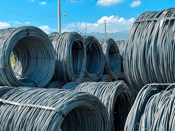 Large coils of steel cables are stacked together outdoors. The cables are neatly wound and secured with metal bands. Power lines and a clear blue sky with scattered clouds are visible in the background.