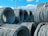 Large coils of steel cables are stacked together outdoors. The cables are neatly wound and secured with metal bands. Power lines and a clear blue sky with scattered clouds are visible in the background.