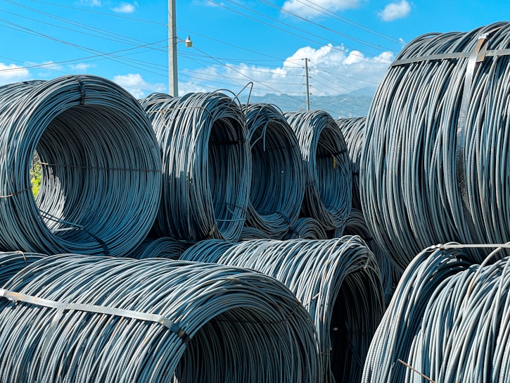 Large coils of steel cables are stacked together outdoors. The cables are neatly wound and secured with metal bands. Power lines and a clear blue sky with scattered clouds are visible in the background.