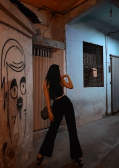 A person strikes a pose in a dimly lit urban alleyway next to a wall featuring graffiti of a face. The scene is characterized by muted lighting and a sense of urban grit.