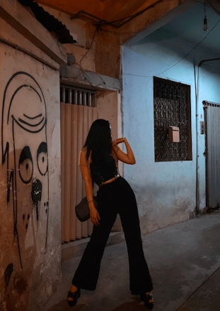 A person strikes a pose in a dimly lit urban alleyway next to a wall featuring graffiti of a face. The scene is characterized by muted lighting and a sense of urban grit.