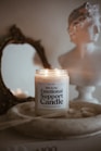 A lit candle labeled 'Emotional Support Candle' sits in a shallow wooden dish. Behind it, a classic-style bust and an ornate, antique mirror add to the aesthetic. The background features soft, neutral tones creating a cozy and serene atmosphere.
