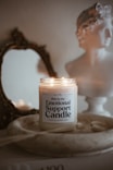 A lit candle labeled 'Emotional Support Candle' sits in a shallow wooden dish. Behind it, a classic-style bust and an ornate, antique mirror add to the aesthetic. The background features soft, neutral tones creating a cozy and serene atmosphere.