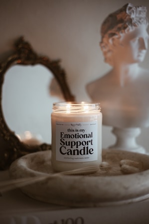 A lit candle labeled 'Emotional Support Candle' sits in a shallow wooden dish. Behind it, a classic-style bust and an ornate, antique mirror add to the aesthetic. The background features soft, neutral tones creating a cozy and serene atmosphere.