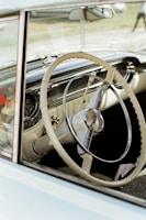 A vintage car's interior is visible, focusing mainly on the intricately designed steering wheel with a chrome and ivory aesthetic. The dashboard features vintage dials and gauges. The scene has an overall nostalgic and classic feel.