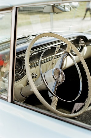 A vintage car's interior is visible, focusing mainly on the intricately designed steering wheel with a chrome and ivory aesthetic. The dashboard features vintage dials and gauges. The scene has an overall nostalgic and classic feel.