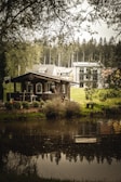 A small wooden cabin is situated near a tranquil pond, surrounded by lush greenery and trees. In the background, modern houses are visible, contrasting with the rustic cabin. The scene is framed by overhanging branches, adding to the natural, serene atmosphere.
