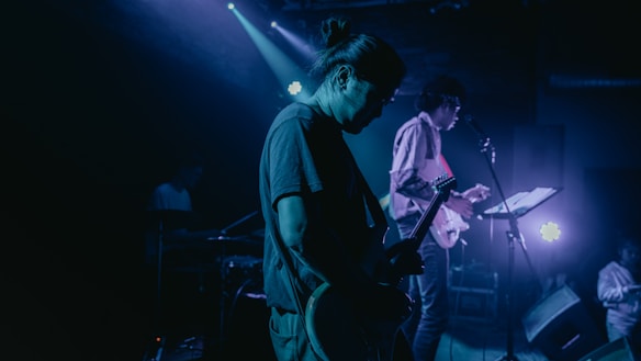 A dimly lit concert scene with musicians performing on stage. The main focus is on a guitarist in the foreground, silhouetted against the stage lights. Another musician is visible in the background, holding a microphone, and a drummer is partially obscured by darkness. The atmosphere is enhanced by blue and purple lighting, creating an intimate and moody setting.