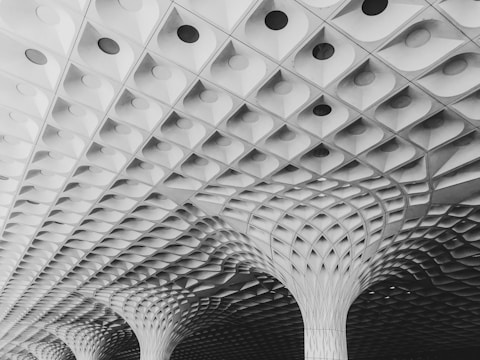 A modern architectural ceiling design featuring intricate geometric patterns and columns that create a visually captivating and symmetrical structure. The ceiling is composed of repetitive, diamond-shaped elements with circular indents, converging towards a series of columns.