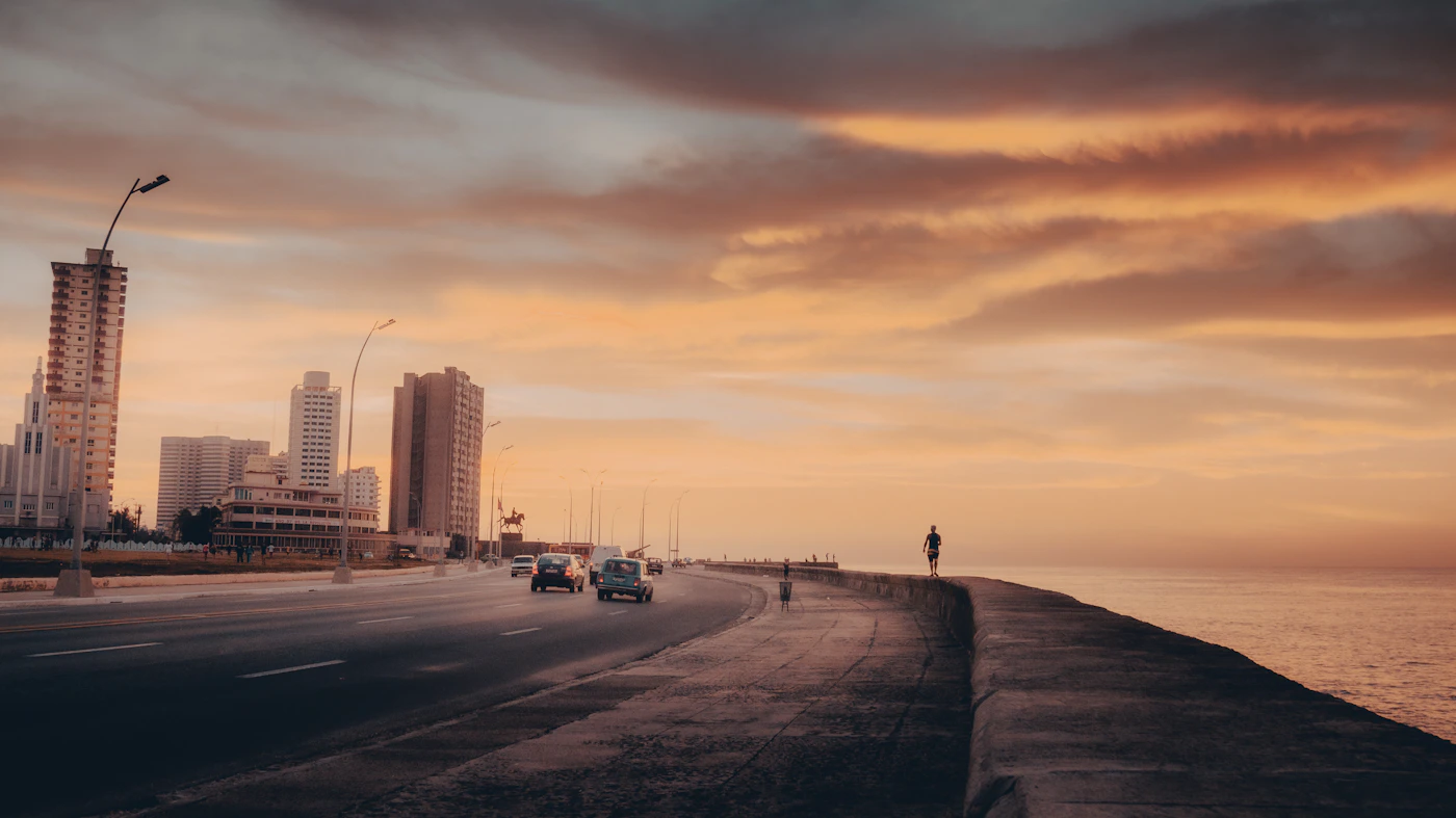 A man standing alone on the Malecon sea wall in Havana Cuba with the grey sea and overcast sky behind him and the aged buildings of the Malecon waterfront stretching behind in the soft Cuban light
