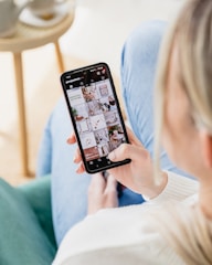 A person is holding a smartphone displaying a social media app with various images. The focus is on the screen, showing an array of lifestyle photos and home decor. The background is slightly blurred, indicating an indoor setting with a chair and table.