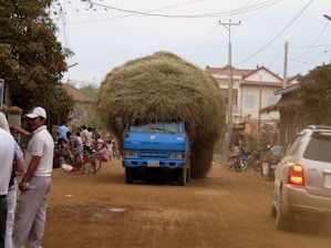 A blue truck heavily laden with dried grass driving on a rural dirt road. The road is surrounded by houses and poles with electric wires. Several people, including a man in a white shirt, are standing on the side of the road next to bicycles and motorbikes.