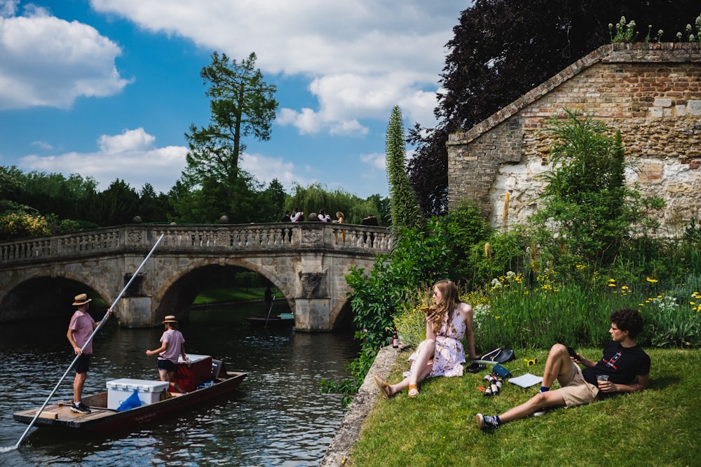 a group of people sitting on the side of a river next to a bridge