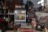 A vintage shop window display features a retro slot machine with the word 'Colossus' on its screen surrounded by various antiques. These include a colorful ceramic vase with floral patterns, a rotary dial telephone, and several decorative figurines. There is also a large old-fashioned map or painting depicting a train and countryside. The reflection of nearby buildings and a parked car can be seen on the window glass.