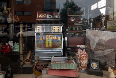 A vintage shop window display features a retro slot machine with the word 'Colossus' on its screen surrounded by various antiques. These include a colorful ceramic vase with floral patterns, a rotary dial telephone, and several decorative figurines. There is also a large old-fashioned map or painting depicting a train and countryside. The reflection of nearby buildings and a parked car can be seen on the window glass.