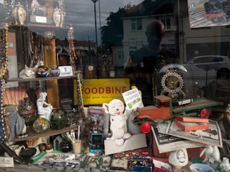 A shop window display is filled with a variety of vintage and eclectic items. There are vases, beaded necklaces, a model airplane, a decorative clock under a glass dome, and several figurines. A large 'Woodbine' advertisement suggests a historical theme, while other signs and objects add a nostalgic feel. The reflection of the street outside, including buildings and cars, is visible on the glass.