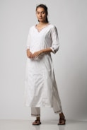 A person is standing wearing a traditional white kurta and trousers, with intricate embroidery on the neckline and cuffs. The outfit is complemented by brown sandals. The individual has long hair tied back and is wearing sunglasses on top of their head.