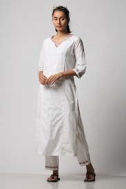 A person is standing wearing a traditional white kurta and trousers, with intricate embroidery on the neckline and cuffs. The outfit is complemented by brown sandals. The individual has long hair tied back and is wearing sunglasses on top of their head.
