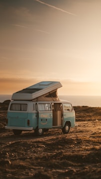 A vintage blue camper van with an elevated roof is parked on a rocky landscape near the ocean at sunset. The open door reveals the interior, and the sky is filled with warm hues.