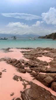 A tranquil beach scene with unique pink sand and dark jagged rocks in the foreground. The turquoise sea is calm, with a few boats gently floating. In the background, there is a distant view of lush green hills under a partly cloudy sky.