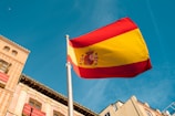 A large, vibrant Spanish flag waving on a flagpole against the backdrop of a clear blue sky. Below, part of a building with neoclassical architectural elements, including arched windows and detailed brickwork, is visible.