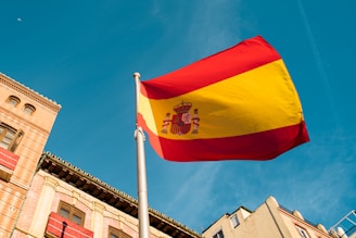 A large, vibrant Spanish flag waving on a flagpole against the backdrop of a clear blue sky. Below, part of a building with neoclassical architectural elements, including arched windows and detailed brickwork, is visible.