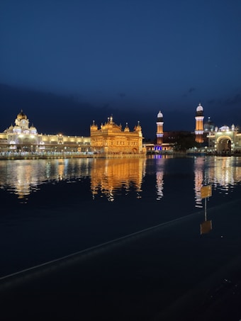 An illuminated temple reflecting in a calm body of water during nighttime. The structure, adorned with glowing golden lights, stands majestically against a dark blue sky. Surrounding buildings are also gently lit, adding to the serene ambiance.