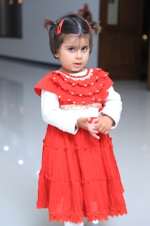 A young child with dark hair tied in pigtails adorned with red clips wears a bright red dress adorned with ruffles and white decorative elements. The dress contrasts with the smooth and shiny light-colored floor beneath. The background is blurred with a hint of festive decor.