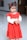 A young child with dark hair tied in pigtails adorned with red clips wears a bright red dress adorned with ruffles and white decorative elements. The dress contrasts with the smooth and shiny light-colored floor beneath. The background is blurred with a hint of festive decor.