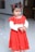A young child with dark hair tied in pigtails adorned with red clips wears a bright red dress adorned with ruffles and white decorative elements. The dress contrasts with the smooth and shiny light-colored floor beneath. The background is blurred with a hint of festive decor.