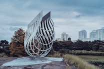 A modern art sculpture made of interwoven metal pieces is set in an outdoor environment with a cityscape in the background. The sculpture features a fluid, wave-like design and stands on a flat, paved area. Surrounding it are green trees and grass, leading to a collection of high-rise buildings under a cloudy sky.