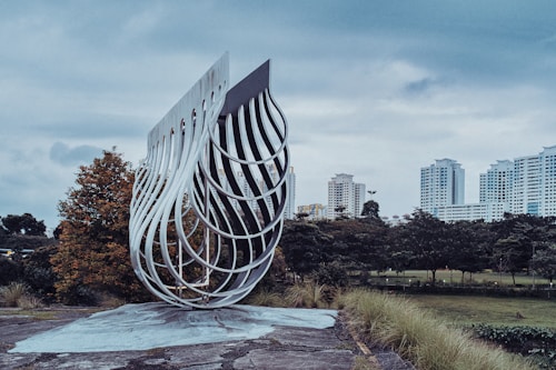 A modern art sculpture made of interwoven metal pieces is set in an outdoor environment with a cityscape in the background. The sculpture features a fluid, wave-like design and stands on a flat, paved area. Surrounding it are green trees and grass, leading to a collection of high-rise buildings under a cloudy sky.