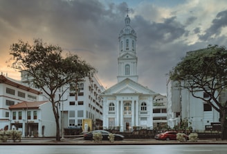 A historic church building stands prominently in the center, flanked by trees and modern urban structures. The sky above is filled with dramatic clouds at sunset, creating a rich contrast of colors.